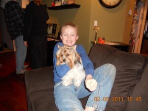 A boy sitting on a couch hugs a small dog in a living room. Two people stand in the background.