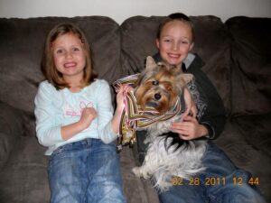 Two children sitting on a couch hold a dog decorated with multiple medals around its neck. The date on the image reads February 28, 2011.