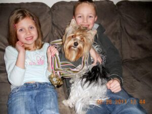 Two children sitting on a couch, holding a dog with multiple medals hanging around its neck. Date stamp reads 02-28-2011 12:44.