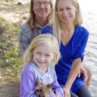 A smiling family with a young girl and a small dog pose together on a lakeside path surrounded by trees.