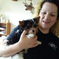 A woman smiles while holding a small black, white, and tan dog indoors. A mounted deer head is visible on the wall behind them.