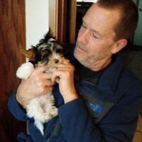 A man in a blue jacket holds a small, fluffy puppy in his left arm while they sit indoors.